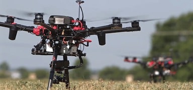 A close-up of a black and red drone hovering above a grassy field, with another similar drone visible in the blurred background. The sky is clear and light blue.