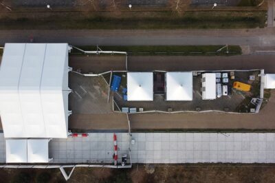 Aerial view of a temporary event site with large white tents, smaller tents, and various equipment arranged along a paved area, surrounded by fencing and walkways.
