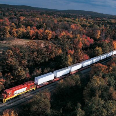 A freight train with a red and yellow locomotive pulls white cargo containers through a dense forest with autumn foliage, featuring vibrant red, orange, and yellow trees under a partly cloudy sky.