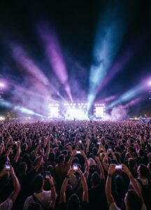 A large crowd at an outdoor concert faces a brightly lit stage, with colorful lights and smoke beams illuminating the night sky. Many people in the audience hold up their phones to capture the performance.