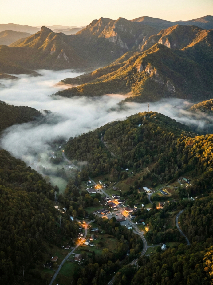 Aerial view of a small village nestled in green, forested mountains at sunrise, with mist and clouds floating in the valleys and warm sunlight illuminating rooftops and winding roads.