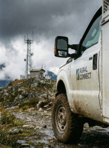 A muddy Rural Connect utility truck is parked on a rocky dirt road near a hilltop communication tower and small building, under a cloudy sky.