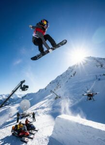 A snowboarder performs a high jump trick in the air above a snowy mountain, with film crews, cameras, and a drone capturing the action under a bright sun and clear blue sky.