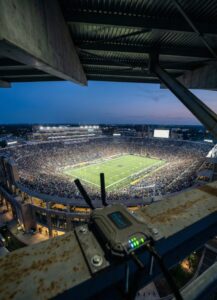 A packed football stadium is illuminated at dusk. The field is brightly lit, and the stands are filled with fans. In the foreground, a small electronic device with antennas and glowing lights is mounted on a railing.