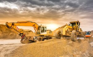Excavator and grader working on a construction site with large dirt mounds, under a dramatic, cloudy sky at sunset.