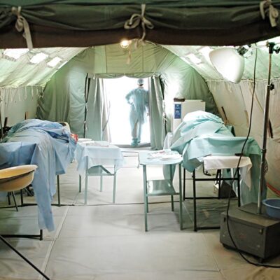 Interior of a medical tent with two patient beds covered in blue sheets, medical equipment, and a person in scrubs standing in the entrance at the far end, light streaming in from outside.