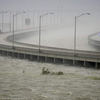 A large, curved bridge over turbulent, flooded water during a storm; heavy rain reduces visibility, and a police vehicle with flashing lights is seen on the otherwise empty bridge.