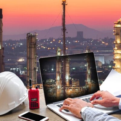 A person types on a laptop at a desk with a hard hat, walkie-talkie, phone, and blueprints, overlooking an industrial refinery at sunset. Tall towers and city buildings are visible in the background.