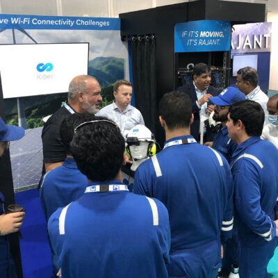 A group of men in blue uniforms gather around a man demonstrating a white helmet at a technology booth, with a screen displaying "Kiber" and signs about Wi-Fi connectivity challenges in the background.