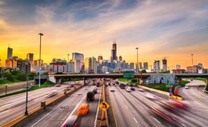 A busy highway with blurred cars in motion leads toward the Chicago skyline at sunset, with the Willis Tower and other skyscrapers visible under a colorful sky.