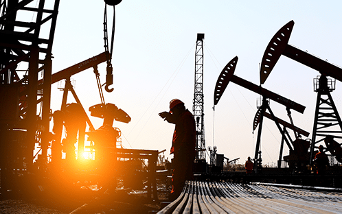 Silhouettes of workers and oil pumpjacks operate at an oilfield during sunset, with the sun low on the horizon and industrial equipment visible in the background.