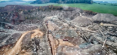 Aerial view of a large open-pit mining site with surrounding dirt roads, equipment, and green fields in the background. A digital network pattern is overlaid on the image.