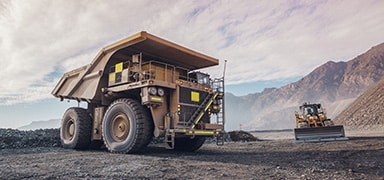 A large mining dump truck is parked on a gravel surface at an open-pit mining site, with mountains in the background and a bulldozer working nearby under a cloudy sky.