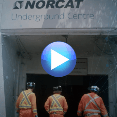 Three workers wearing orange safety gear and helmets enter the NORCAT Underground Centre through a white entrance, with a large play button overlayed at the center of the image.