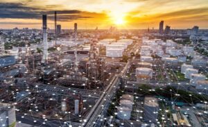 Aerial view of an industrial oil refinery complex at sunset, with storage tanks, pipelines, and city buildings in the background. Digital network graphics overlay the foreground, symbolizing connectivity and technology integration.
