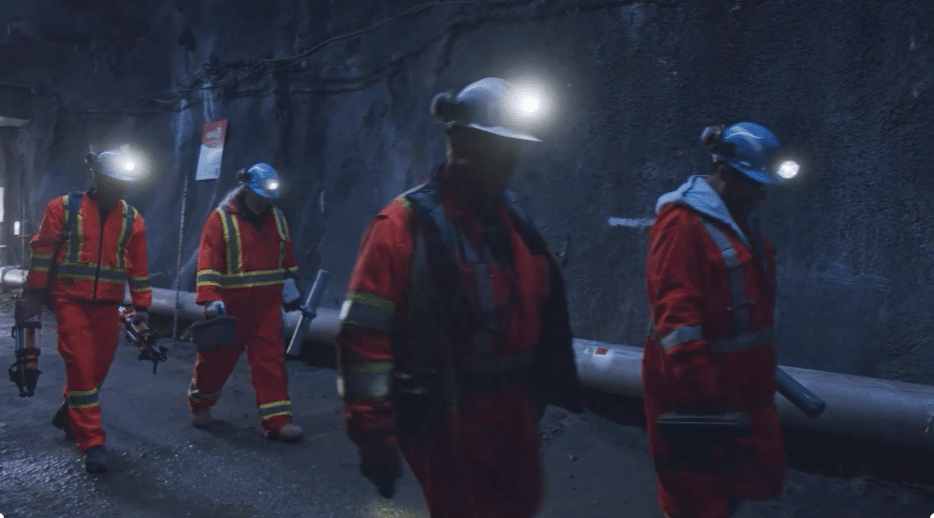 Four workers wearing orange safety gear and helmets with headlamps walk through a dimly lit underground tunnel, carrying equipment. The tunnel walls are dark and rough, with pipes running along one side.