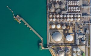 Aerial view of an industrial facility with large storage tanks and silos beside turquoise water, featuring a long pier extending from the shore into the water.