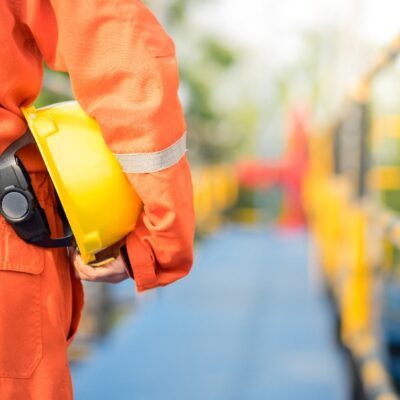 A worker in an orange safety uniform holds a yellow hard hat while standing on a walkway with safety railings, suggesting a construction or industrial site. The background is blurred.
