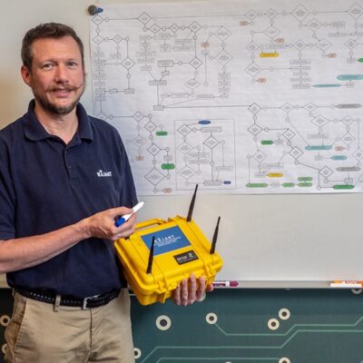 A man stands in front of a whiteboard with a complex flowchart, holding a yellow device with two antennas and a blue label in one hand, and a whiteboard marker in the other.