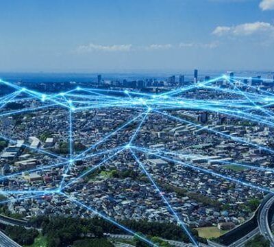 A cityscape with digital blue lines and nodes overlayed, representing connectivity and network communication across urban infrastructure under a blue sky.