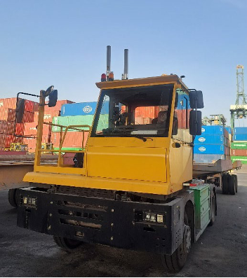 A yellow terminal tractor truck parked at a shipping yard, with stacked shipping containers in the background under a clear blue sky.