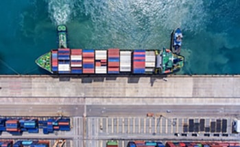 Aerial view of a large cargo ship docked at a port, loaded with colorful shipping containers. Trucks and more containers are visible on the dock beside the ship.