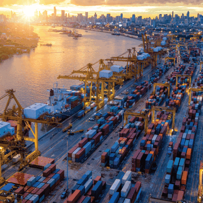 Aerial view of a busy shipping port at sunset, with large cargo ships docked and numerous colorful shipping containers stacked along the docks, yellow cranes, and a city skyline in the background.