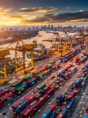 Aerial view of a busy shipping port at sunset with colorful cargo containers, large cranes, and a river. The city skyline is visible in the background, and digital network lines overlay the scene, symbolizing smart technology.