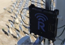 A black device with a large blue "R" and a wireless signal icon is mounted on a pole outdoors. In the background, rows of small, round, reflective solar panels are arranged on sandy ground.