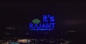 A nighttime sky display features illuminated drones forming the words "It's RAJANT" in blue and green lights above a dark landscape with scattered lights below.