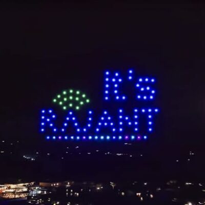 A nighttime sky display features illuminated drones forming the words "It's RAJANT" in blue and green lights above a dark landscape with scattered lights below.