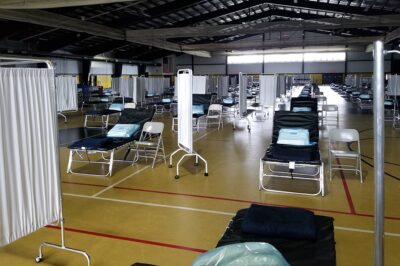 A large gymnasium is set up as a temporary medical facility, with rows of cots, chairs, privacy screens, and folded blankets, all arranged in an organized manner under fluorescent lighting.