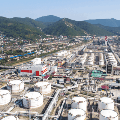 Aerial view of an industrial oil refinery complex with large storage tanks, pipelines, and various buildings, set near a town with green mountains in the background.