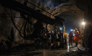 Two workers in orange safety gear and helmets stand near heavy machinery inside a dimly lit, rocky tunnel with wet, muddy ground. Bright lights illuminate parts of the tunnel wall and the workers.