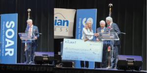 A group of people stands on a stage with SOAR banners and a large ceremonial check for $875,000 addressed to an education foundation. A man speaks at a podium while others shake hands and smile.