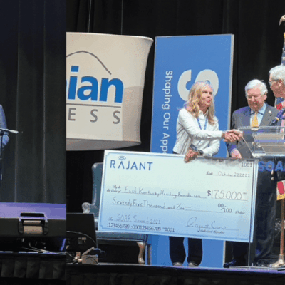 A group of people stands on a stage with SOAR banners and a large ceremonial check for $875,000 addressed to an education foundation. A man speaks at a podium while others shake hands and smile.