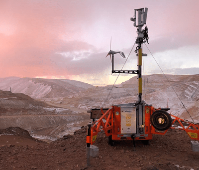 A portable weather station with solar panels and a wind turbine stands on rocky terrain with snow-dusted hills and a colorful sky in the background.