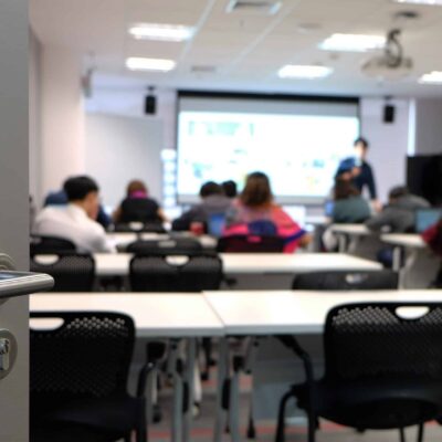A classroom seen from an open doorway, with students seated at desks facing a screen and a standing instructor at the front. The room is brightly lit with modern furniture.