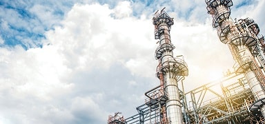 Industrial refinery with tall metal towers and complex piping against a cloudy sky, sunlight illuminating part of the structure.