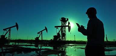 A silhouette of a person wearing a hard hat checks a tablet near oil pumpjacks at sunset, with reflections visible in water on the ground.