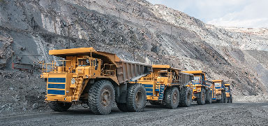 Four large yellow mining trucks are parked in a row on a gravel road in an open-pit mine, with steep rocky walls in the background.