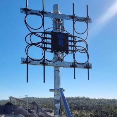 A metal antenna tower with multiple cables and rectangular frames stands outdoors on a sunny day, overlooking a landscape with greenery and industrial structures in the background.