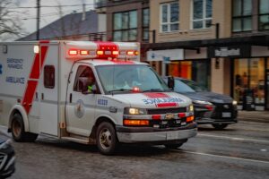 An ambulance with flashing lights drives down a wet city street, passing by parked cars and storefronts on a cloudy day.