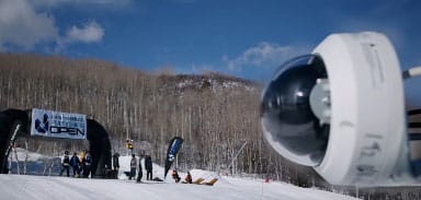 A security camera is mounted in the foreground overlooking a snowy ski event, with people, banners, and leafless trees visible in the background under a blue sky.