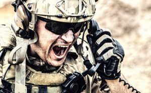 A soldier wearing a helmet, sunglasses, and tactical gear yells into a field phone, appearing intense and urgent, with a blurred outdoor background.