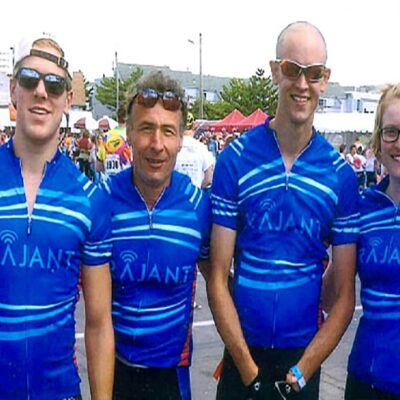 Six cyclists in matching blue "Rajant" jerseys stand together smiling at an outdoor event, with other participants and tents visible in the background. One person wears a race bib numbered 7443.