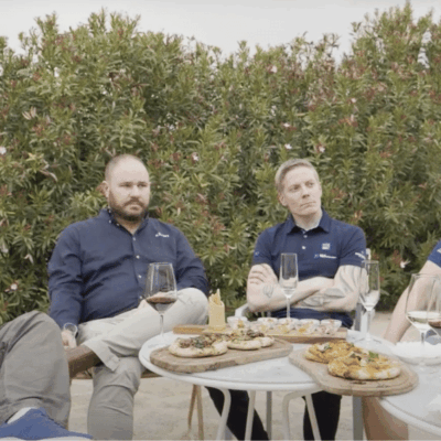 Five men in matching navy shirts are seated outdoors around a table with wine glasses and plates of appetizers. Green bushes are in the background, and the group appears to be engaged in conversation.