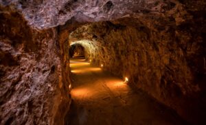 A dimly lit underground tunnel with rough, rocky walls and a dirt floor, illuminated by small lights placed along the pathway, creating a warm glow through the cavern.