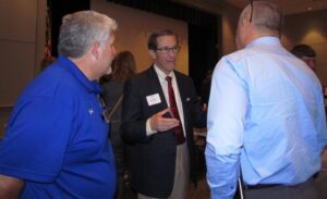 Three men stand in conversation at an indoor event. One man, wearing a suit and nametag, gestures as he speaks, while the other two, dressed in casual shirts, listen attentively. Other people and chairs are visible in the background.
