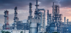 A large industrial oil refinery with numerous tall metal towers, pipes, and tanks, set against a cloudy sky at dusk. Greenery is visible at the bottom of the image.